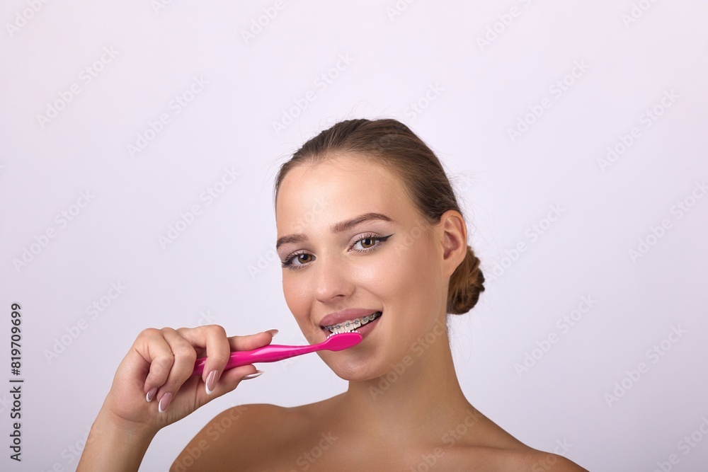 A young woman with a captivating smile brushing her teeth with a pink toothbrush. Her brown hair stands out against a white background, emphasizing her oral care routine. A young woman with a captivating smile brushing her teeth with a pink toothbrush. Her brown hair stands out against a white background, emphasizing her oral care routine.