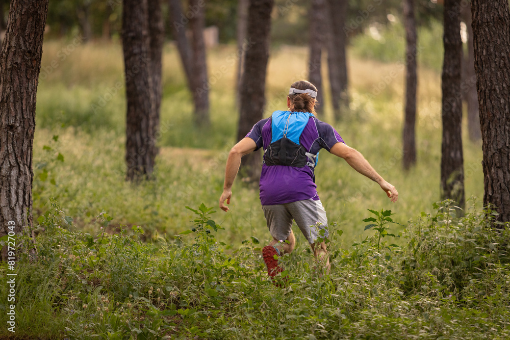 A man is running through a forest with a backpack on. The man is wearing a purple shirt and gray shorts A man is running through a forest with a backpack on. The man is wearing a purple shirt and gray shorts