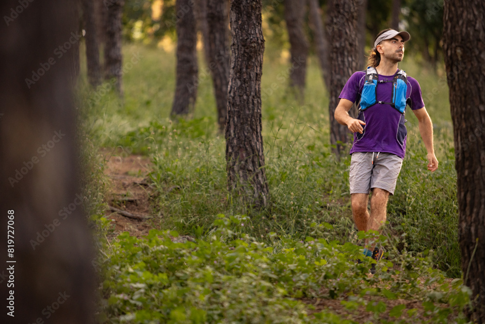 A man in a purple shirt and gray shorts is walking through a forest. He is carrying a backpack and a water bottle