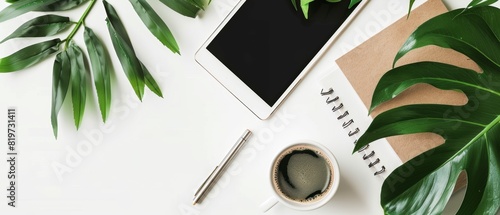 Top view of a tidy workspace with a notepad, coffee cup, tablet, and green leaves on a white background, perfect for business visuals