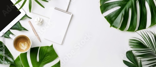 Top view of a clean workspace with a notepad, coffee cup, tablet, and green leaves on a white background, ideal for business visuals photo