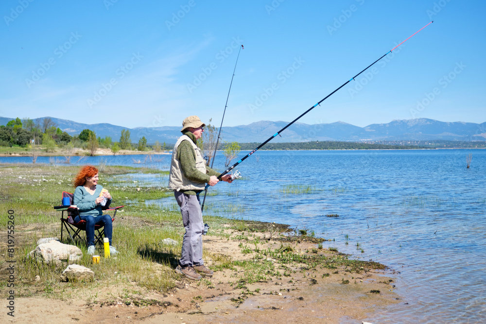 A man and a woman are fishing in a lake. The man is holding a fishing rod and the woman is sitting in a chair eating a sandwich. Scene is peaceful and relaxing A man and a woman are fishing in a lake. The man is holding a fishing rod and the woman is sitting in a chair eating a sandwich. Scene is peaceful and relaxing