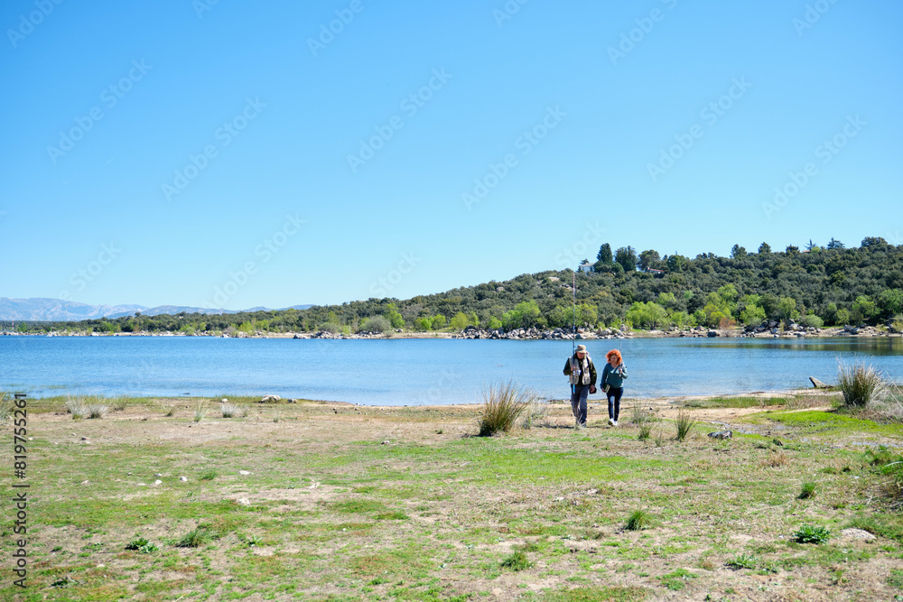 A couple is walking by a lake after fishing. The sky is clear and the water is calm