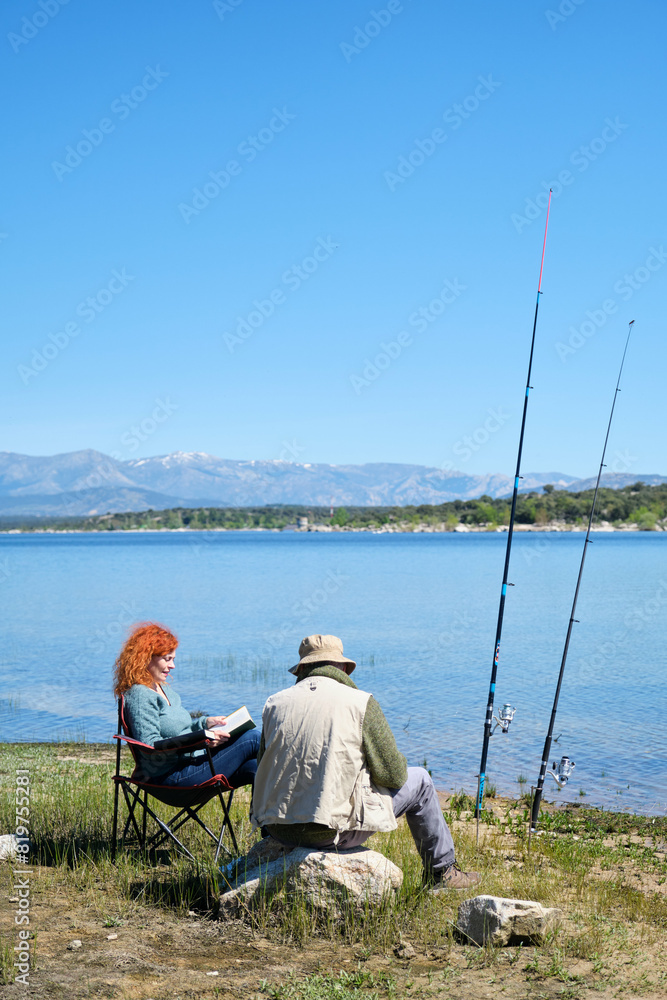A man and a woman are sitting by a lake, fishing. The man is looking at the fishing rod and the woman is reading a book. The scene is peaceful and relaxing A man and a woman are sitting by a lake, fishing. The man is looking at the fishing rod and the woman is reading a book. The scene is peaceful and relaxing
