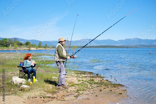 A man and a woman are fishing in a lake. The man is holding a fishing rod and the woman is sitting in a chair eating a sandwich. Scene is peaceful and relaxing