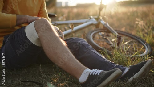 Unrecognizable man wraps bandage around knee after receiving injury. Cyclist administers first aid to himself after falling and injuring knee in rural environment. Fixing joint with tight bandage. photo