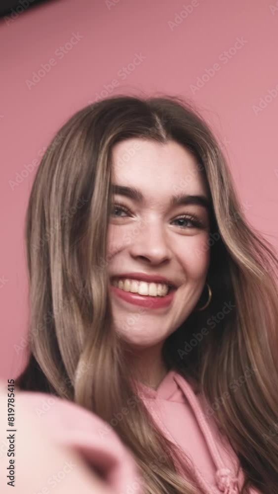 Real time handheld shot of charming smiling young female with long hair and in makeup looking at camera with kissing lips while standing in room with stretched hand against pink backdrop Real time handheld shot of charming smiling young female with long hair and in makeup looking at camera with kissing lips while standing in room with stretched hand against pink backdrop