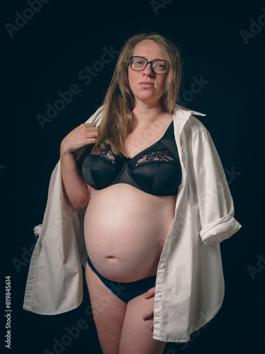 The mom-to-be shows off her pregnant belly against a dark background. She is dressed in a black bra with lace details, blue underwear and a white shirt over her shoulders.