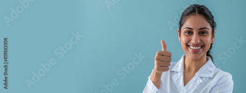 Wallpaper Mural banner, wide shot of young Indian female doctor in white scrubs doing delicious thumbs up sign, smiling looking in camera, happy expression, blue background copy space Torontodigital.ca
