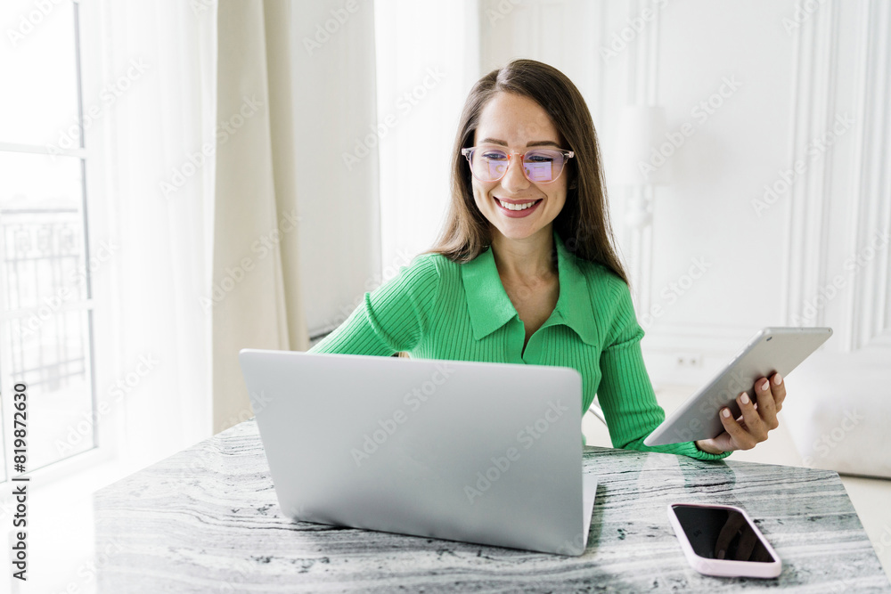 A professional woman with glasses, wearing a green sweater, using a tablet while working on a laptop at a stylish desk. A professional woman with glasses, wearing a green sweater, using a tablet while working on a laptop at a stylish desk.