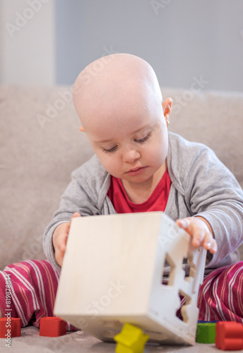 Baby girl with cancer and hair loss due to chemotherapy playing with wood toy on the sofa © bymandesigns