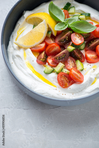 Bowl of greek feta dip topped with fresh tomatoes and cucumber, vertical shot on a light-grey granite surface, middle closeup