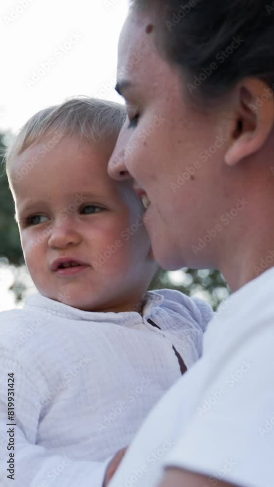 Mom carries little son in arms walking against lush tree branches at bright sunlight. Woman and toddler explore nature of countryside closeup slow motion Vertical Shot. Mom carries little son in arms walking against lush tree branches at bright sunlight. Woman and toddler explore nature of countryside closeup slow motion Vertical Shot.