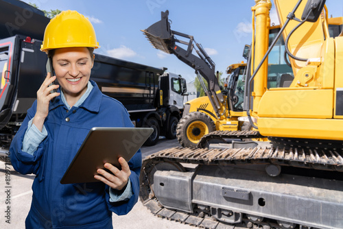 Female engineer in a helmet with a digital tablet stands next to construction excavators.