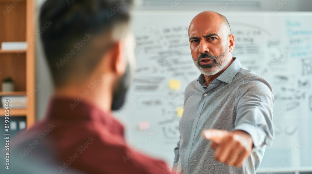 A man with a beard and gray hair wearing a gray shirt pointing ata whiteboard with a stern expression engaged in a discussion with another man whose back is turned to the camera.
