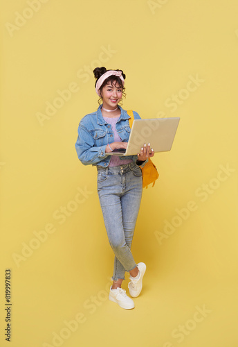 Portrait of happy smiling young Asian college student with laptop and backpack ready to school isolated on yellow background.