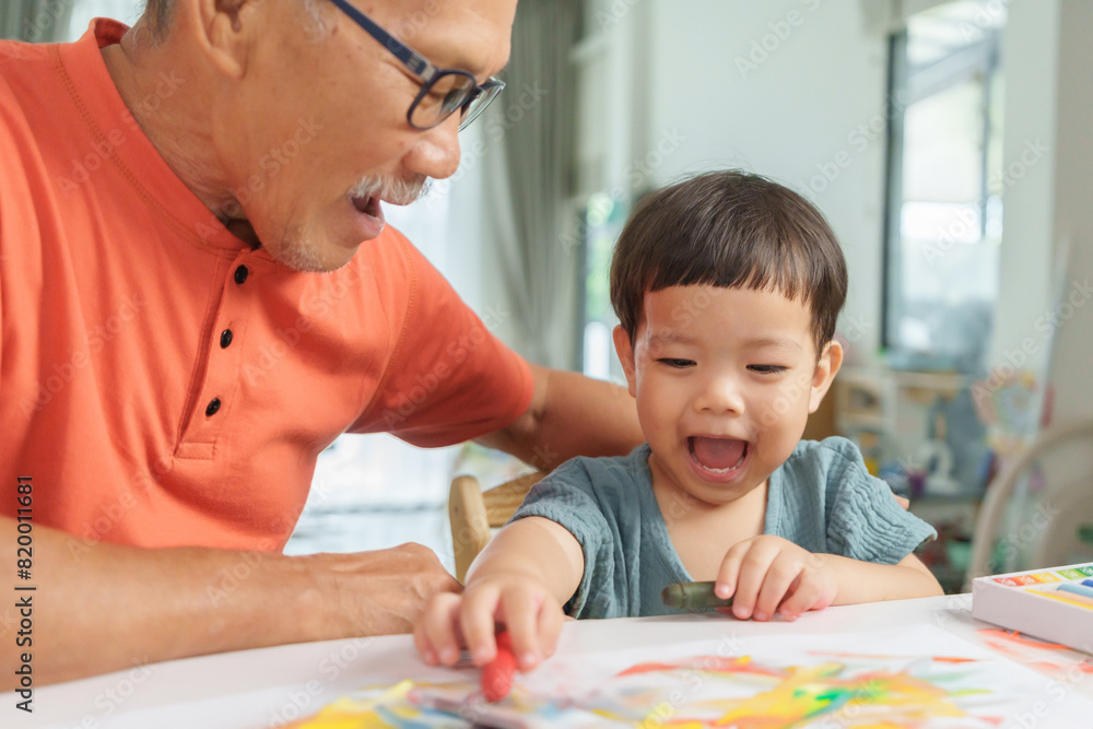 Asian Senior man and Grandson or young boy laughing and drawing together at home, showcasing creativity and happiness in a shared activity. Asian Senior man and Grandson or young boy laughing and drawing together at home, showcasing creativity and happiness in a shared activity.