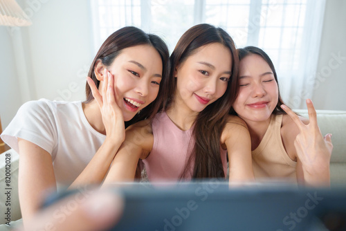 Three young Asian women smiling and making peace signs while taking a selfie with smartphone on a couch, capturing a joyful and friendly moment indoors.