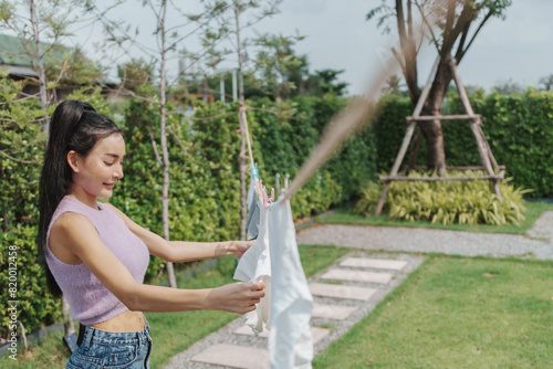 Smiling Asian Woman hanging clothes on a clothesline in a sunny backyard, showing a serene and peaceful daily chore in a green garden.