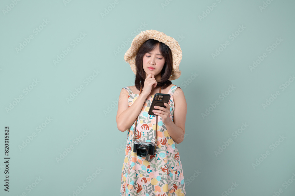 Thinking on vacation concept. Happy Asian woman wearing casual summer dress with hat and camera thinking gesture while using mobile phone isolated on pastel green background. Thinking on vacation concept. Happy Asian woman wearing casual summer dress with hat and camera thinking gesture while using mobile phone isolated on pastel green background.