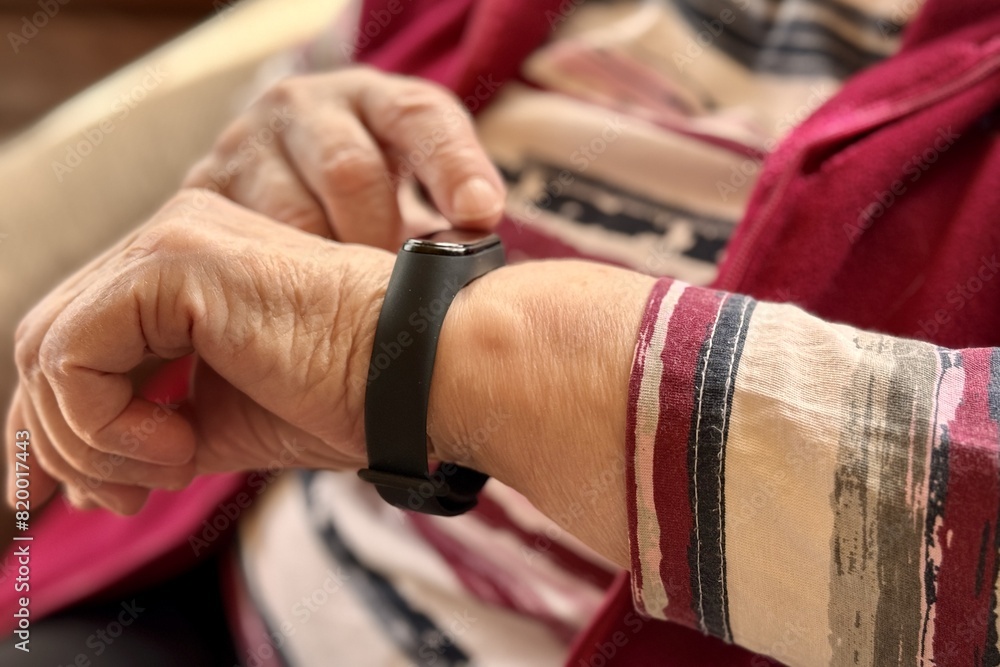 The black smart band with display touchpad and heart rate monitor. The old woman sitting in armchair touches and looks at the wristband of pulse monitor indoors. Fitness tracker