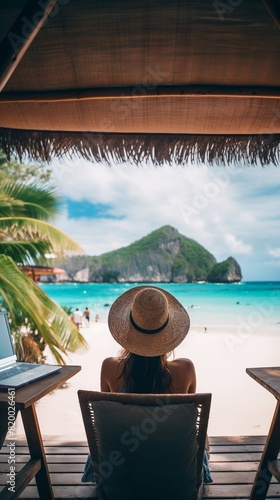 A person in a hat relaxes on a beach chair under a thatched roof  facing the turquoise ocean and scenic mountains.