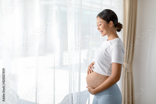 Happy pregnant woman in white shirt and blue pants holding belly, standing near window with curtain, smiling and looking down, indoor natural light. Free Download Happy pregnant woman in white shirt and blue pants holding belly, standing near window with curtain, smiling and looking down, indoor natural light. - GFXLoad