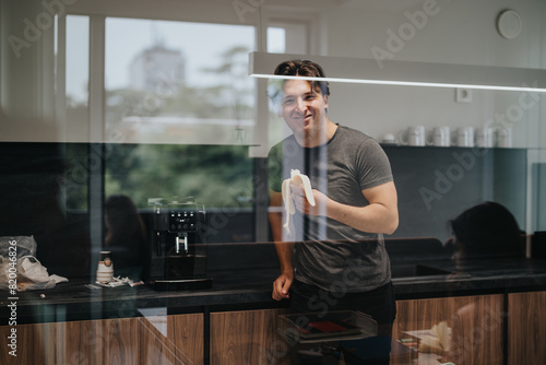 A young man enjoys a quick snack of a banana in the sleek, modern kitchen of his office, portraying a relaxed break during a busy workday.