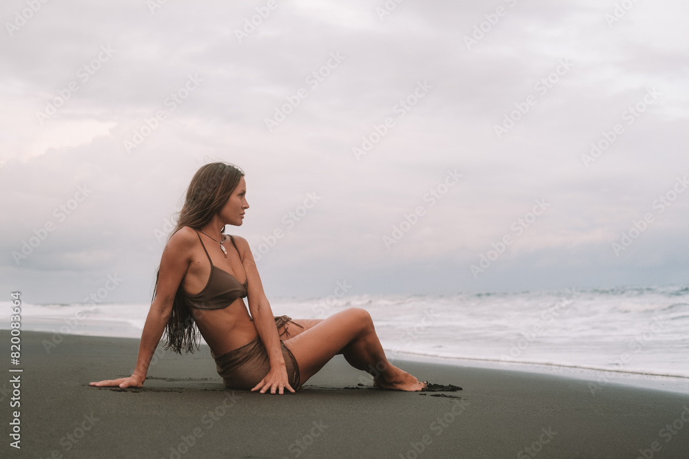 A young sexy woman with long hair sits on a black sand beach in a bikini against the background of the ocean and enjoys her vacation. Summer vacation on a tropical beach with volcanic sand. A young sexy woman with long hair sits on a black sand beach in a bikini against the background of the ocean and enjoys her vacation. Summer vacation on a tropical beach with volcanic sand.