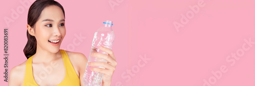 A smiling young woman in a yellow sports bra holding a bottle of water against a pink background. She looks refreshed and energized, highlighting the importance of staying hydrated during workouts.