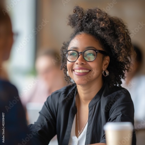 A woman with curly hair and glasses is smiling at the camera. She is wearing a black jacket and white shirt