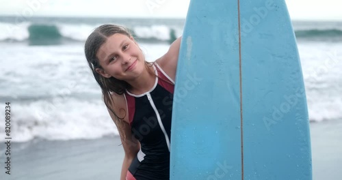 Portrait of a teenage girl who stands on the beach and looks out from behind a blue surfboard and shows a shaka sign with her hand. Summer vacation for a teen, leisure surfing in the ocean, lifestyle. photo