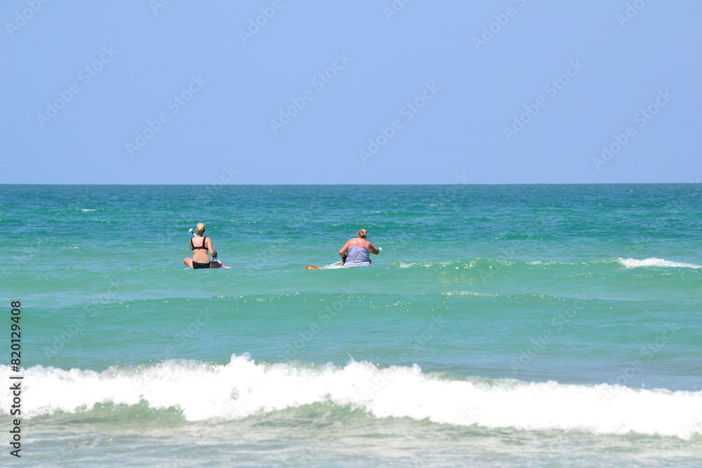 Two women are paddleboarding on the turquoise waters, with waves gently cresting at the shore beneath a clear blue sky at Ponce Inlet, Florida.