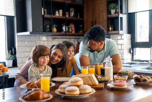 Cheerful couple embracing and talking with children during breakfast at kitchen table at home.