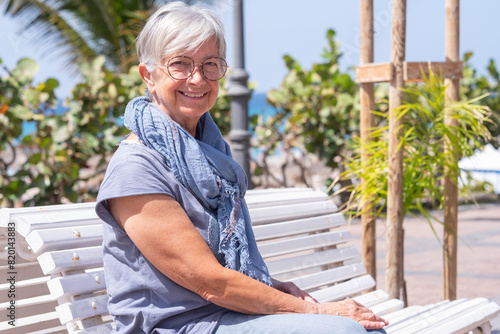 Relaxed white-haired senior woman sitting outdoors on a sunny day near the beach. Portrait of smiling attractive elderly lady enjoying free time and vacation #820143883