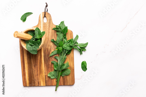 Wooden mortar with fresh green mint leaves on the wooden board on white marble table top view, copy space for your design. Herbal and aromatheraphy concept.
