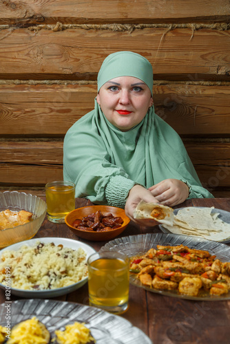 A young Muslim woman in a light khimar at the table takes lamb with her hand during the celebration of Eid al-Fitr