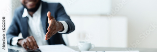 Cropped of african american businessman outstretching his hand towards camera, sitting at desk at modern office, greeting business partner, panorama with copy space photo