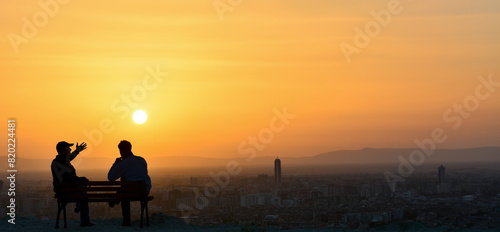 Silhouette of two friends sitting on a park bench and talking during a warm sunset.