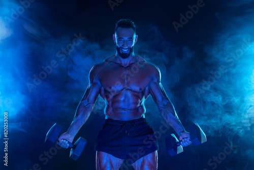 Sporty fit man athlete with dumbbells make fitness exercises on blue neon background. Download cover for music collection for fitness classes. Sports recreation.