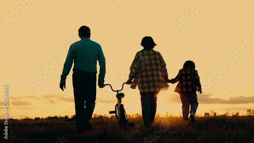 Family silhouette walking at sunset nature field together back view slow motion. Mother father and son with bicycle going holding hands at rural countryside meadow sunrise bright sun sky horizon photo