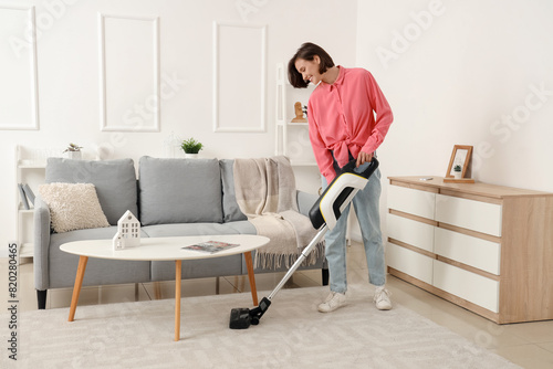Beautiful young woman cleaning carpet with cordless stick vacuum cleaner in living room