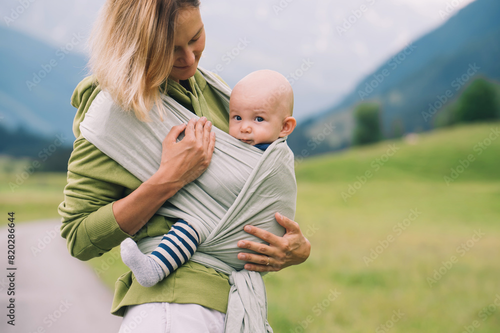 Fototapeta premium Babywearing. Mother and baby on nature outdoors. Baby in wrap carrier. Woman carrying little child in baby sling in green mint color. Concept of green parenting, natural motherhood, postpartum period.