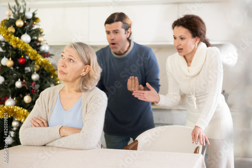 Couple actively scold mother who listens and thinks about it for сhristmas, sitting at table in kitchen near christmas tree © JackF