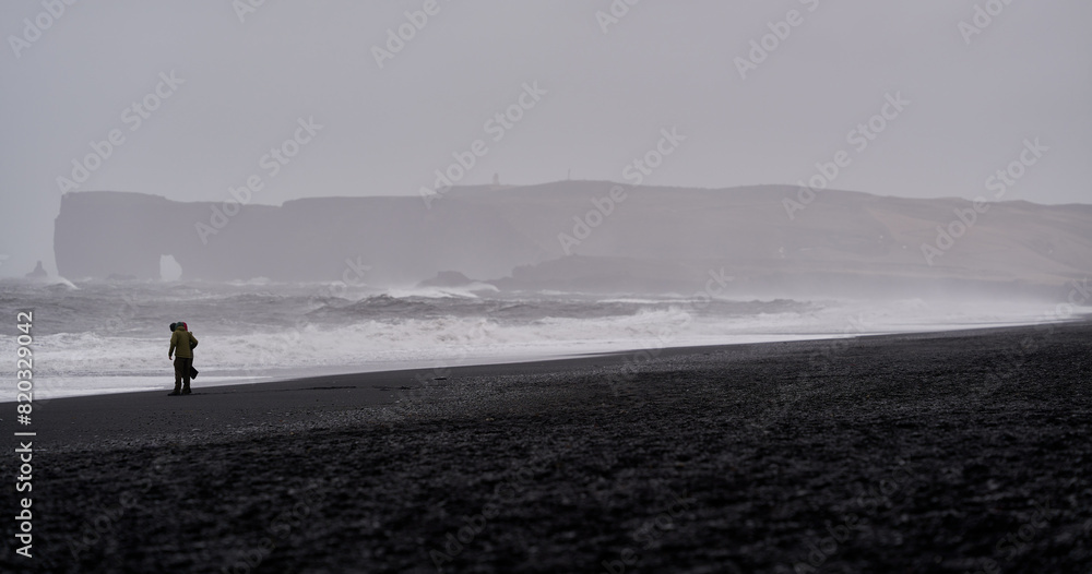 Panoramic view of Dyrhólaey cliffs from the black beach of Reynisfjara in Iceland with 2 unrecognizable tourists admiring the landscape with strong waves