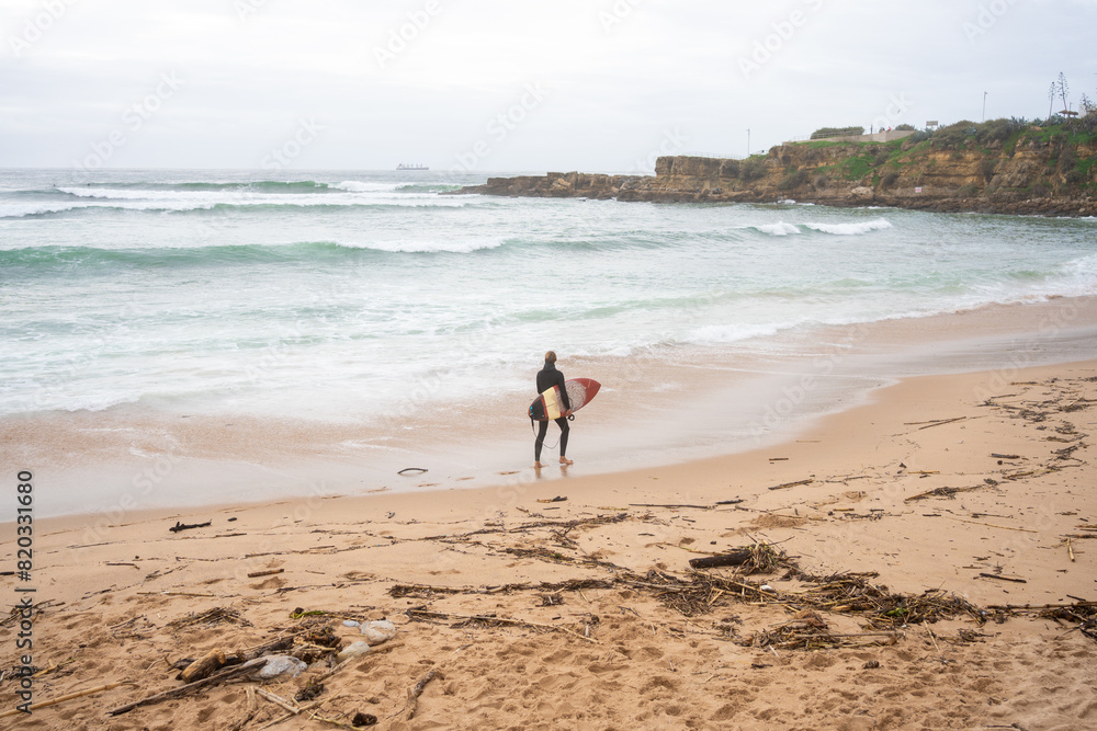 Anonymous surfer carrying surfboard gazing at sea waves rushing towards the shore. Unknown person is ready for surfing session while standing on sandy beach, engaging in extreme water sport. Anonymous surfer carrying surfboard gazing at sea waves rushing towards the shore. Unknown person is ready for surfing session while standing on sandy beach, engaging in extreme water sport.