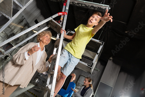 Young boy standing on stepladder and reaching his hand in escape room. His grandmother standing next to him and holding stepladder. photo