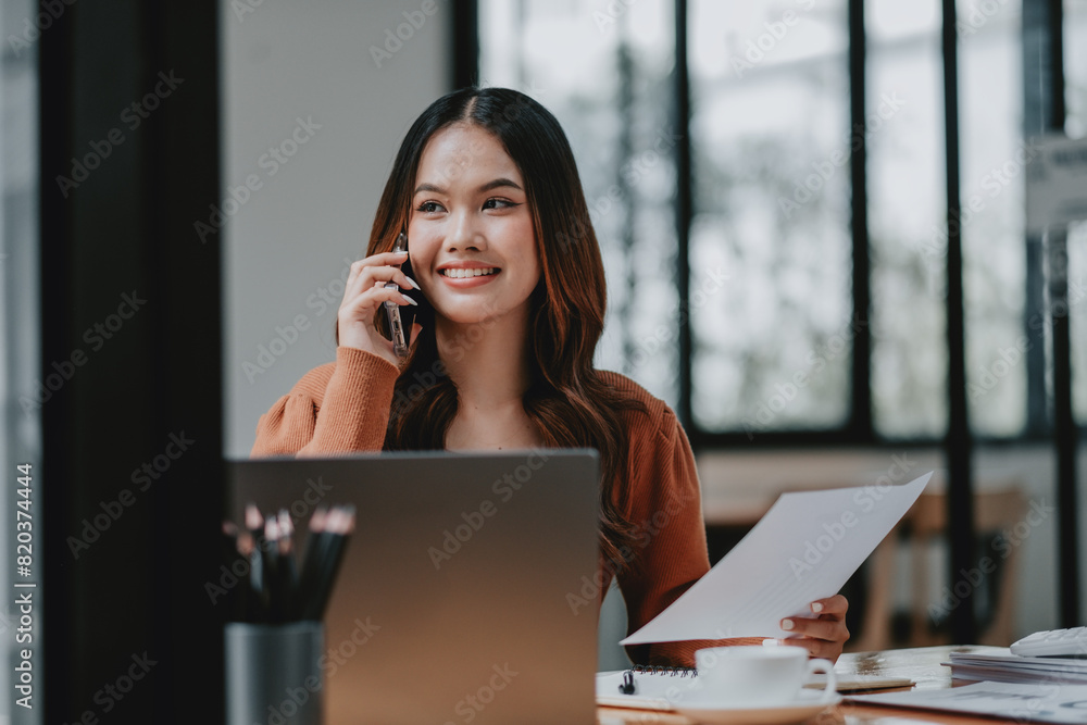 A woman is talking on her cell phone while sitting at a desk with a laptop and a cup. She is smiling and she is enjoying her conversation A woman is talking on her cell phone while sitting at a desk with a laptop and a cup. She is smiling and she is enjoying her conversation