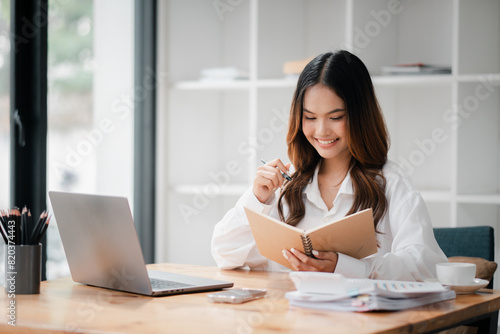A woman is sitting at a desk with a laptop and a notebook. She is smiling and she is enjoying her work