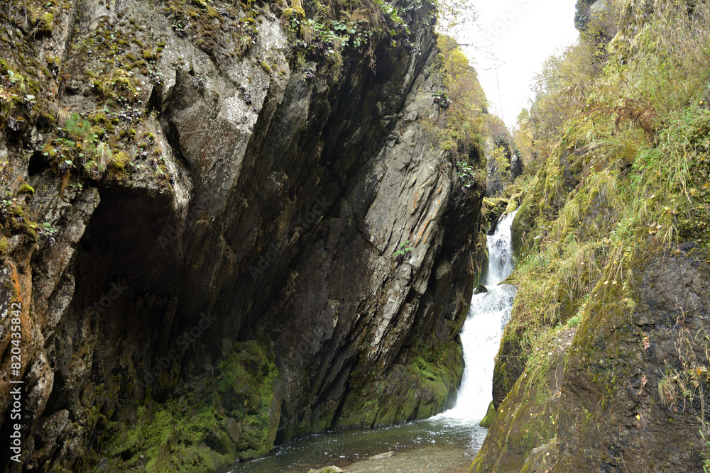 A waterfall sandwiched between two rocks, a stormy stream flows down an autumn forest on a cloudy day. A waterfall sandwiched between two rocks, a stormy stream flows down an autumn forest on a cloudy day.
